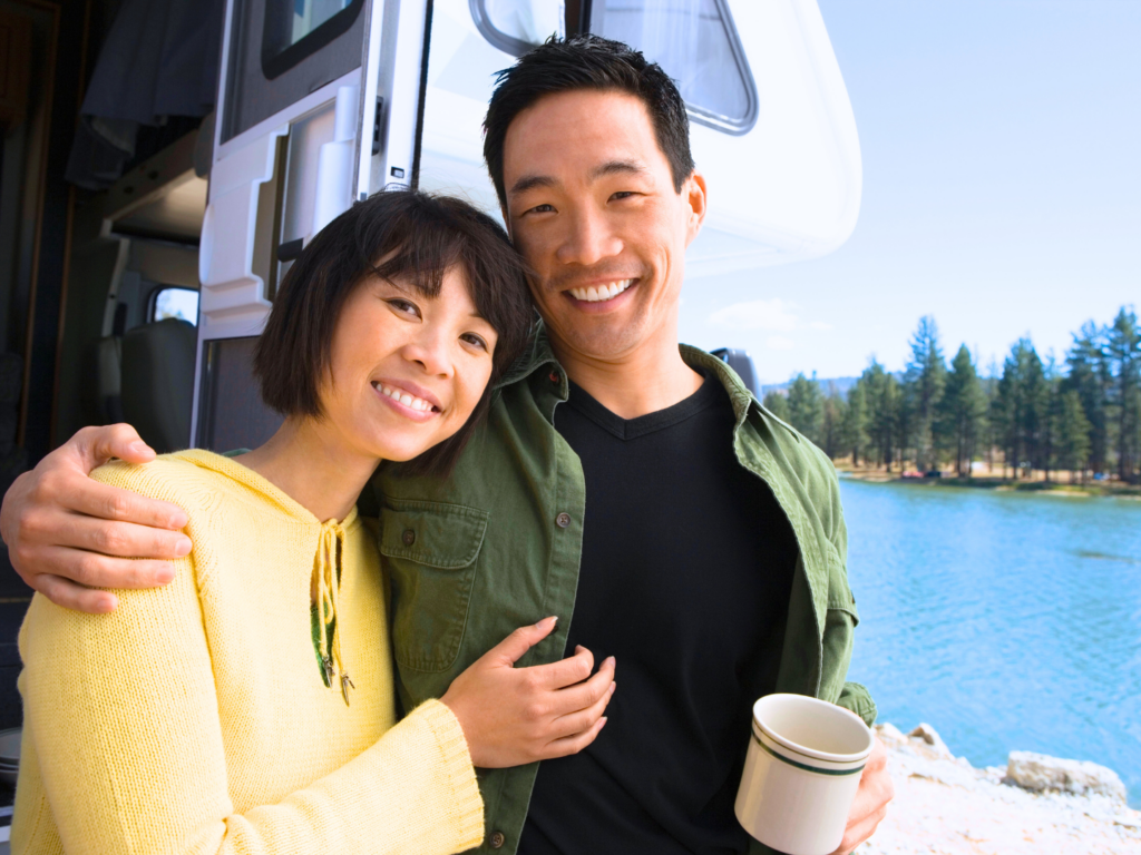 a couple outside of the camper they use to travel around the country in a spot overlooking a lake smiling as they know they used their rv insurance checklist to prepare for this trip